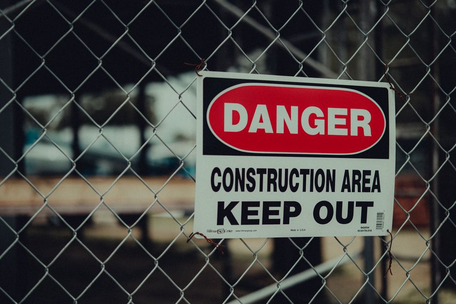 A rectangular sign with a black background and bold red and white lettering reading 'KEEP OUT' is suspended by a thin wire from a weathered wooden plank. The sign is positioned at a slight downward an